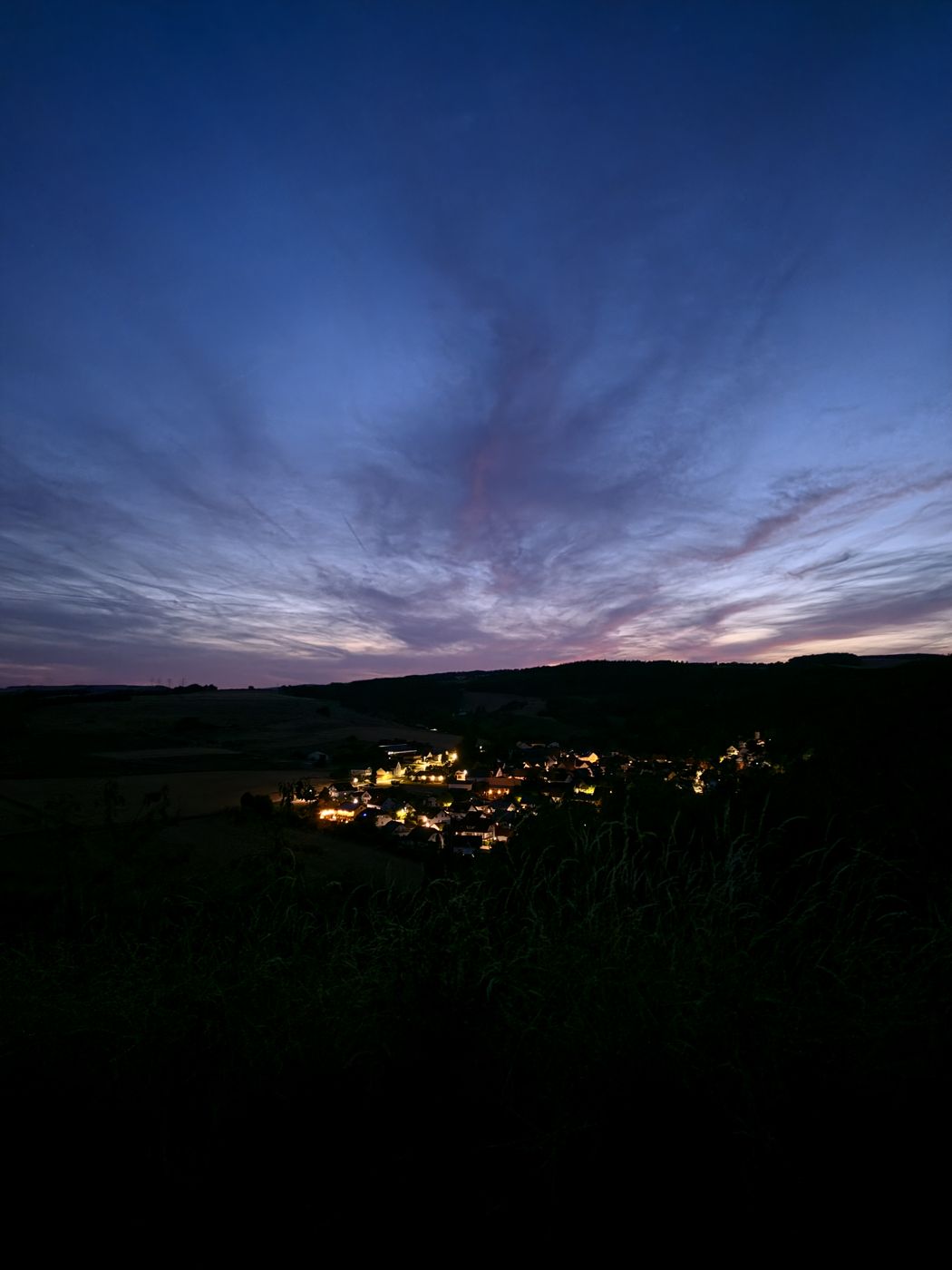 Nachtaufnahme: beleuchtetes Dorf im dunklen Tal, darüber letzte blaue und rosa Dämmerungswolken