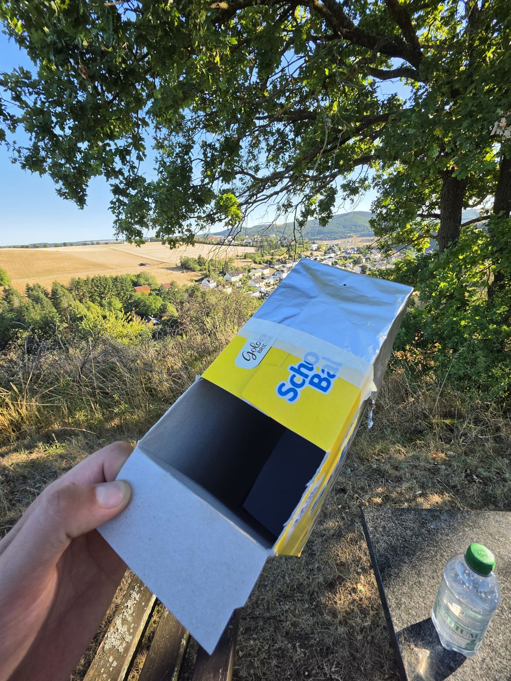 Selbstgebaute Lochkamera aus einer Schoko-Balls-Müslipackung mit Folie und kleinem Loch, in der Hand gehalten, dahinter das Tal