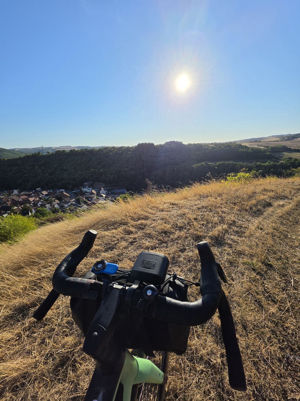 Blick über den Lenker des Gravelbikes auf die tief stehende Sonne über bewaldeten Hügeln, unten im Tal ein Dorf