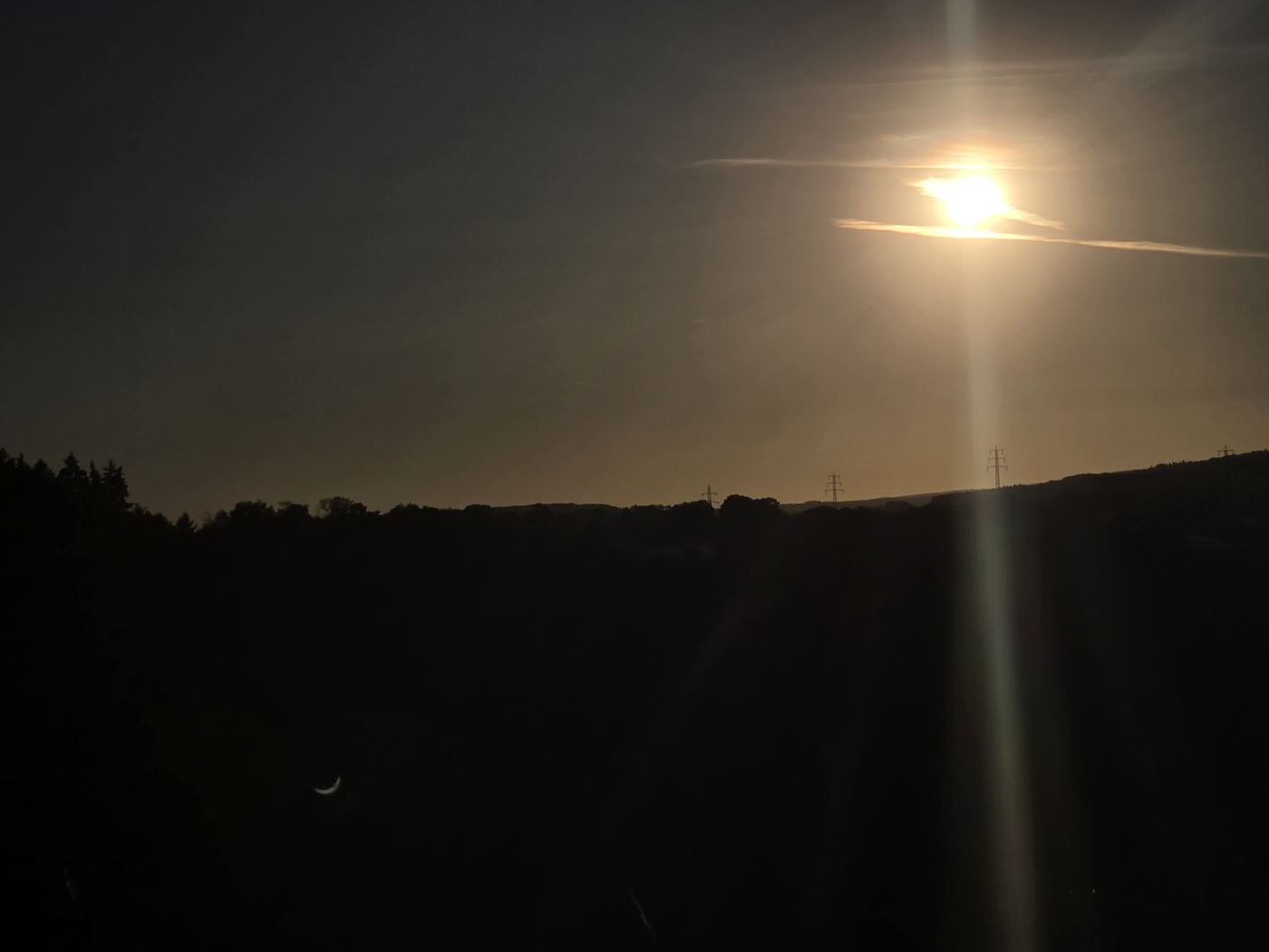 Dunkle Abendstimmung mit tief stehender Sonne hinter Wolkenband, unten links im Bild ein sichelförmiger Linsenreflex