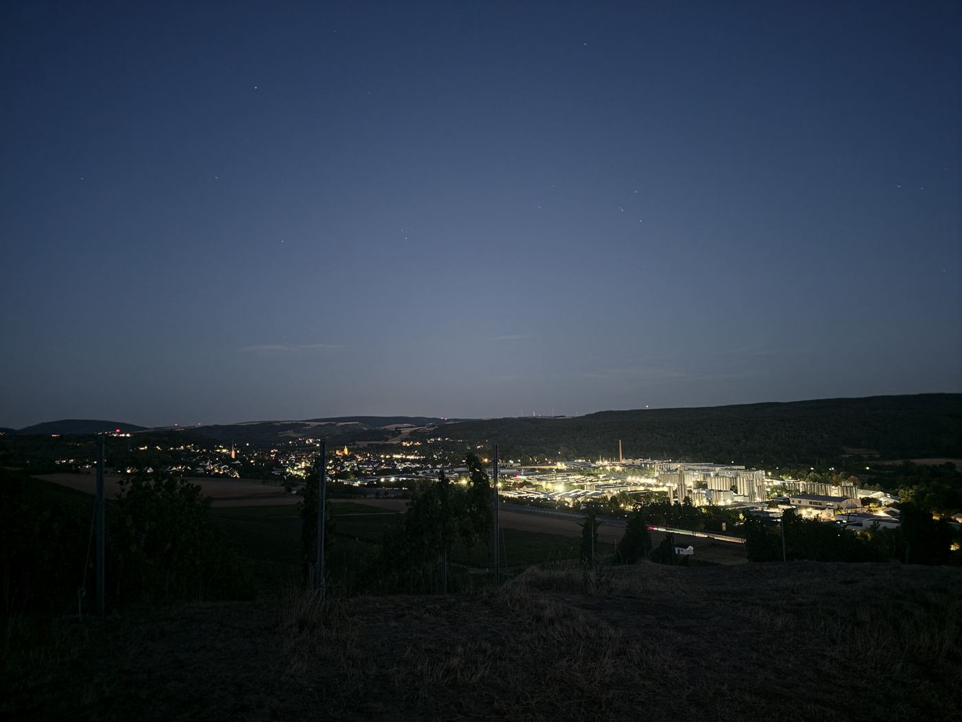 Nachtaufnahme aus den Weinbergen: unten leuchten Dorf und ein hell erleuchtetes Industriegelände im Tal, oben werden die ersten Sterne sichtbar