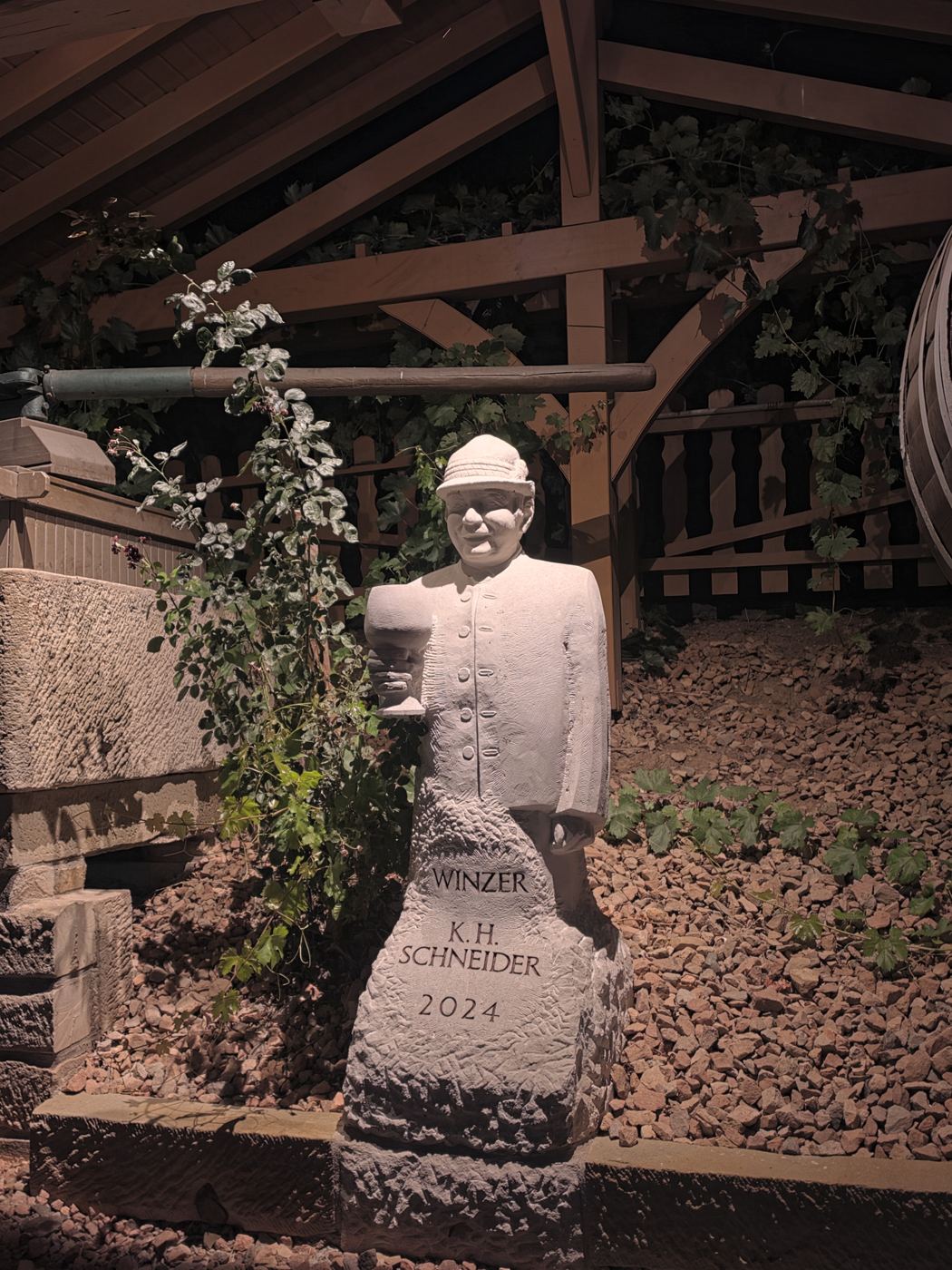 Weiße Steinstatue eines Winzers mit Hut an einem Weingut bei Nacht, umgeben von Reben und einem Holzfass