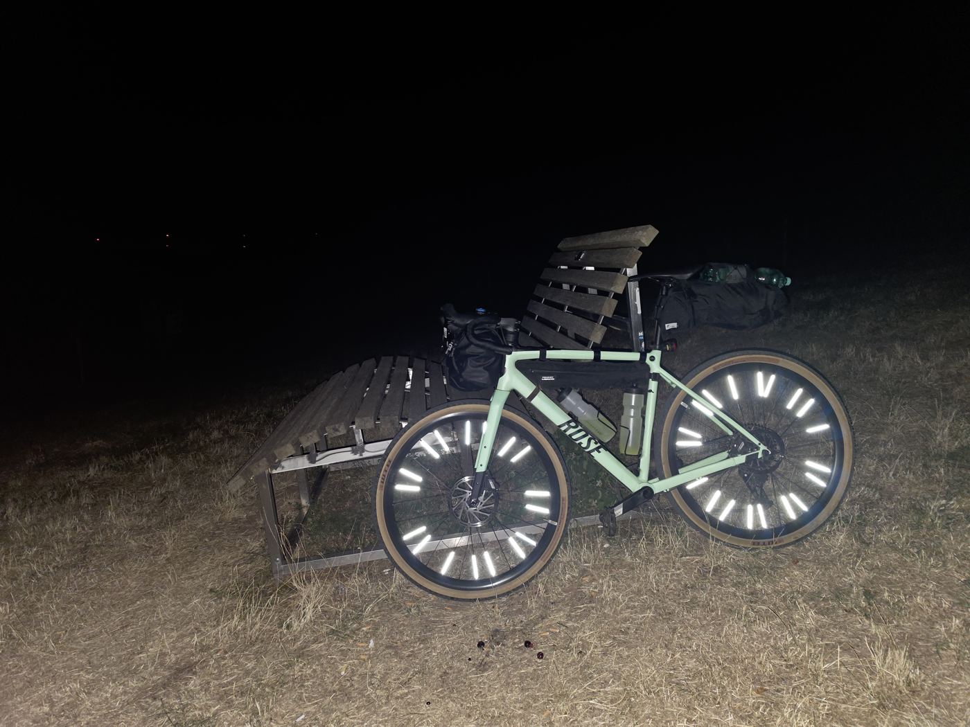 Das Gravelbike mit reflektierenden Speichen lehnt nachts an zwei Holzliegen, auf einer liegt das Gepäck