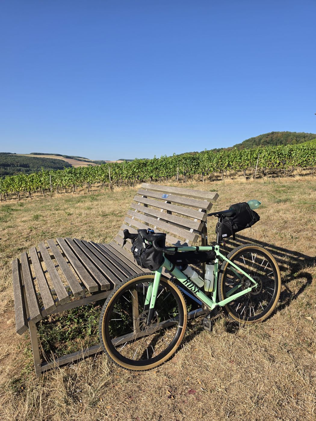 Das komplett bepackte mintgrüne Gravelbike lehnt abfahrbereit an den zwei Holzliegen im Morgenlicht