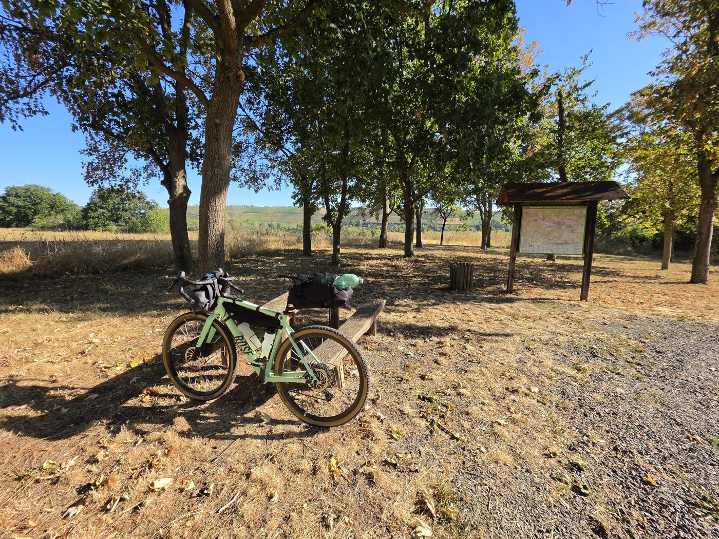 Bepacktes Gravelbike lehnt an einer Holzbank im Baumschatten, rechts eine überdachte Wandertafel, dahinter Weinberge